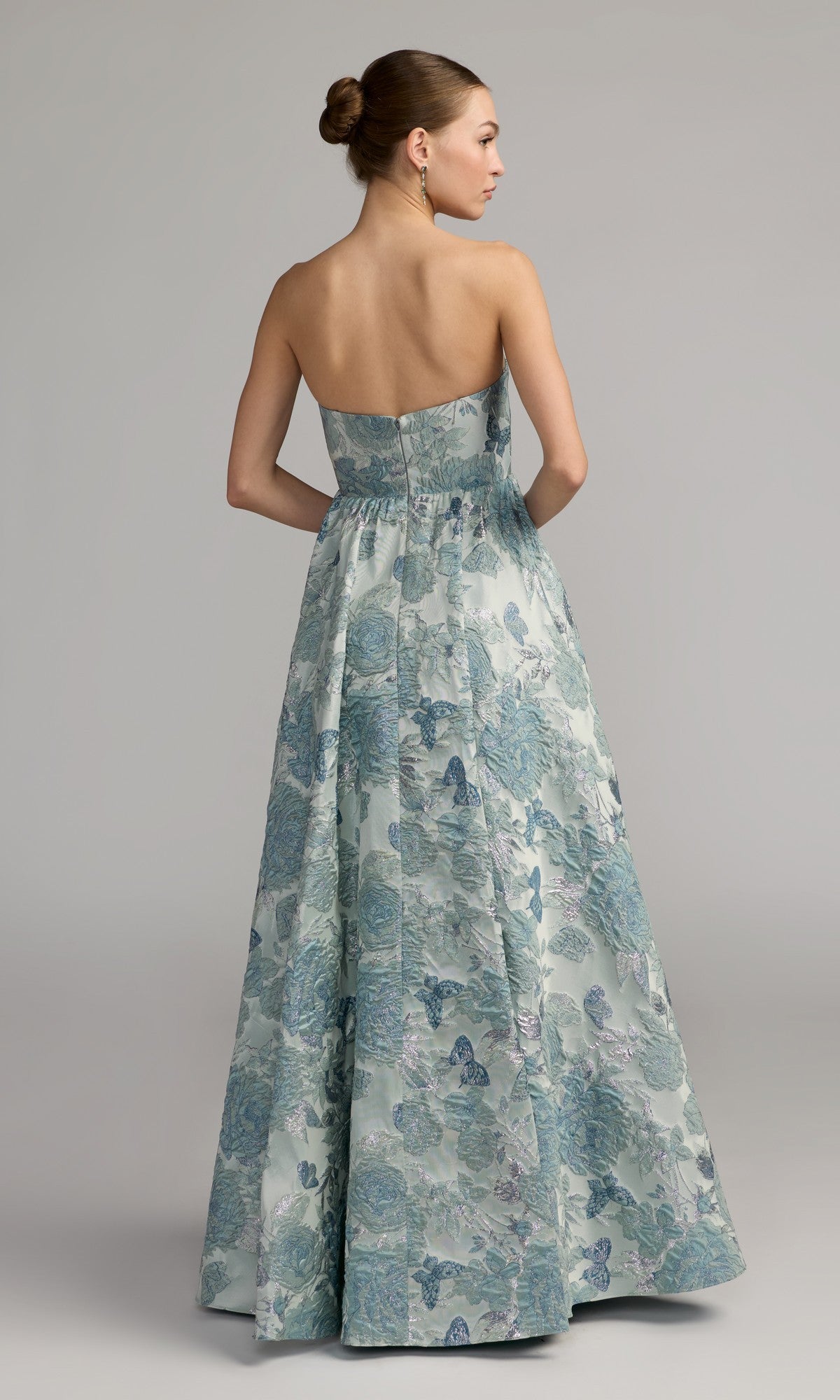A woman stands facing away against a plain gray background, her hair styled in a neat bun, wearing the Mac Duggal 68999 long formal dress—a strapless, floor-length ballgown adorned with elegant blue and silver floral brocade.