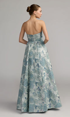 A woman stands facing away against a plain gray background, her hair styled in a neat bun, wearing the Mac Duggal 68999 long formal dress—a strapless, floor-length ballgown adorned with elegant blue and silver floral brocade.