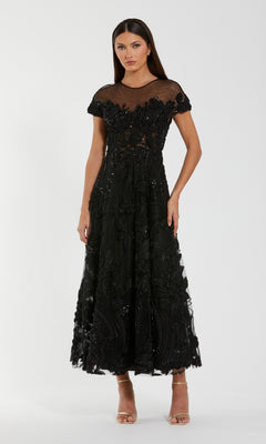 A woman wears the Mac Duggal 69003 Cap-Sleeve Black Formal Dress, a mid-length formal gown with sheer details and floral embellishments, styled with nude open-toe heels.