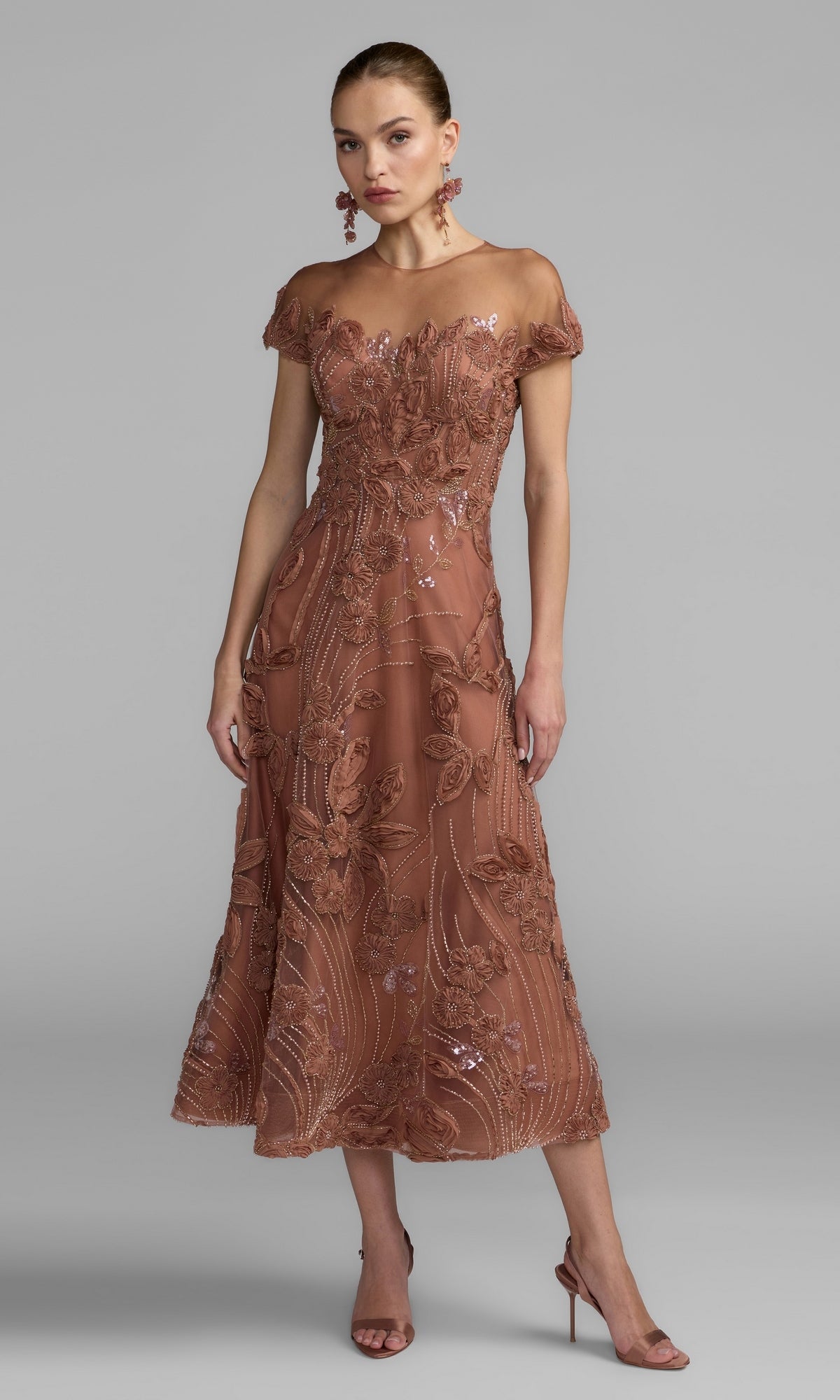 A woman wearing a cap-sleeve, rust-colored formal dress (Mac Duggal 69003) with elegant details, reminiscent of classic evening style, paired with matching earrings and high-heeled sandals, stands against a plain gray background.