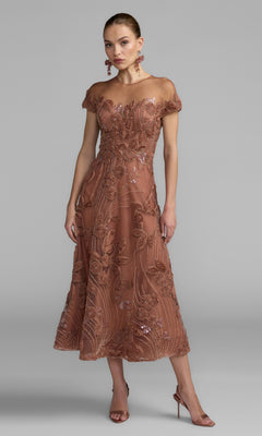 A woman wearing a cap-sleeve, rust-colored formal dress (Mac Duggal 69003) with elegant details, reminiscent of classic evening style, paired with matching earrings and high-heeled sandals, stands against a plain gray background.
