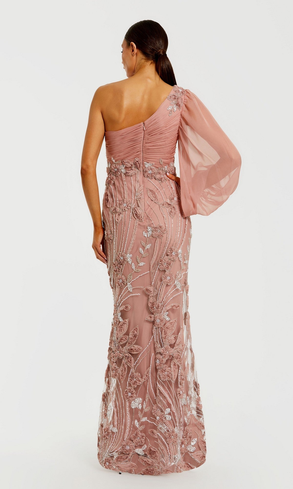 A woman is standing with her back to the camera against a plain white background, wearing the Long Formal Dress: Mac Duggal 69019—an elegant, long pink gown featuring a one-shoulder design, sheer sleeve, and delicate floral detailing.