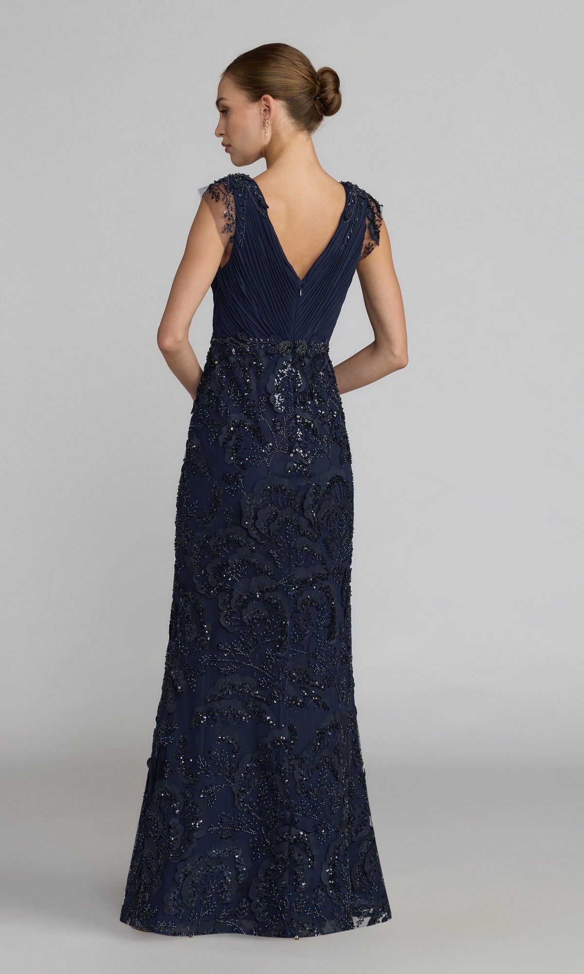 A woman stands facing away, wearing the Mac Duggal 69022 Long Beaded Rose Formal Dress—a floor-length gown in navy blue with cap sleeves, intricate detailing, and a V-shaped back.