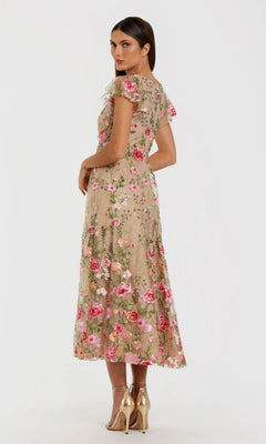 A woman stands in profile against a plain background, wearing the Mac Duggal 69024 Tea-Length Party Dress—a beige lace midi dress with floral embroidery in pink and green, short sleeves, and a flattering v-neckline—paired with gold high-heeled sandals.