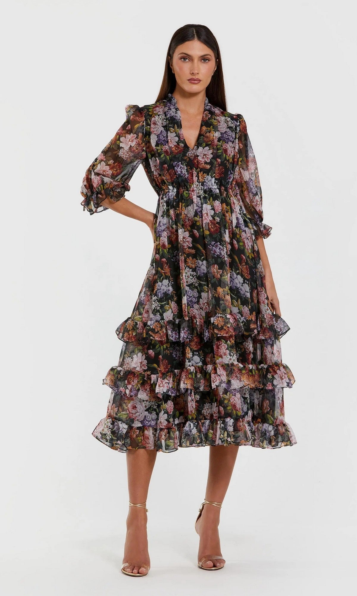 A woman models the Mac Duggal 69031 Midi Floral Wedding-Guest Dress in Black Multi—featuring sheer sleeves, a ruffle tiered skirt, and floral print—paired with strappy heeled sandals against a plain light background.