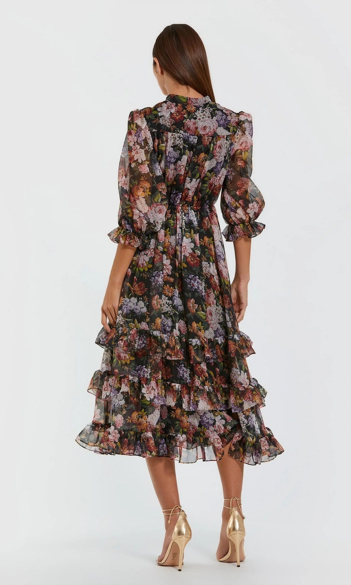 A woman stands facing away, wearing the Mac Duggal 69031 Midi Floral Wedding-Guest Dress—featuring ruffled tiers, long sheer sleeves, and floral chiffon—paired with gold high-heeled shoes against a plain light background.