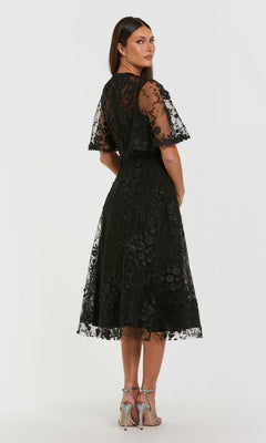 Woman in a Mac Duggal 69036 midi wedding guest dress with floral embroidery and short sheer sleeves, standing in heels and facing slightly away from the camera.