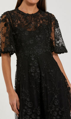 Woman in a black Mac Duggal 69036 Midi Wedding Guest Dress featuring floral embroidery and sheer short sleeves, shown from shoulders to mid-thigh against a plain background.