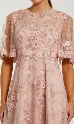 A woman wears the Midi Wedding Guest Dress: Mac Duggal 69036 in light pink, featuring floral lace embroidery and a sheer overlay on the bodice and sleeves.