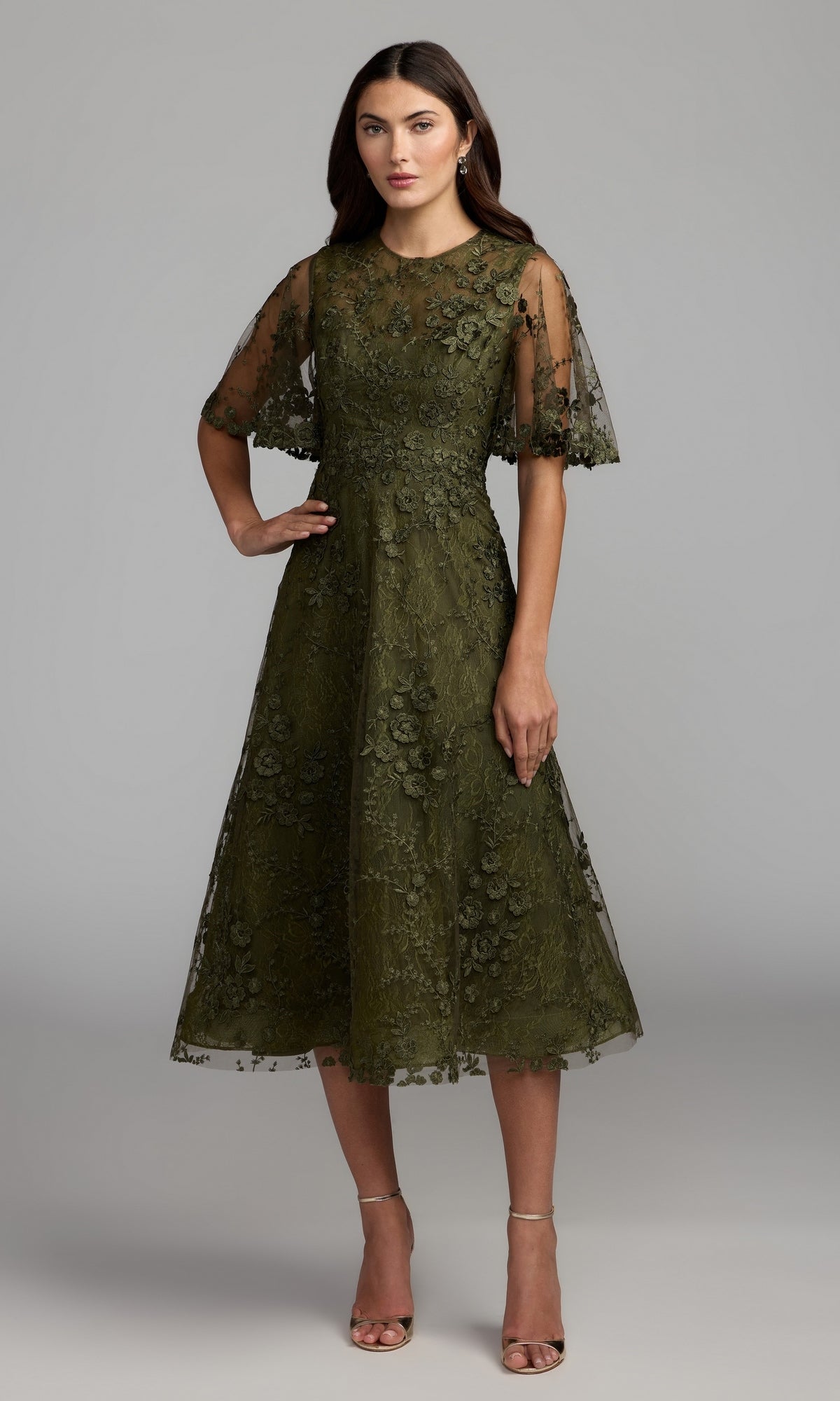 A woman stands in the Lace Midi Wedding Guest Dress: Mac Duggal 69036, featuring a knee-length, olive lace design with delicate floral embroidery and sheer short sleeves. Paired with nude heels against a plain gray background, this chic olive dress is perfect for attending weddings.