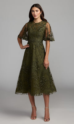 A woman stands in the Lace Midi Wedding Guest Dress: Mac Duggal 69036, featuring a knee-length, olive lace design with delicate floral embroidery and sheer short sleeves. Paired with nude heels against a plain gray background, this chic olive dress is perfect for attending weddings.