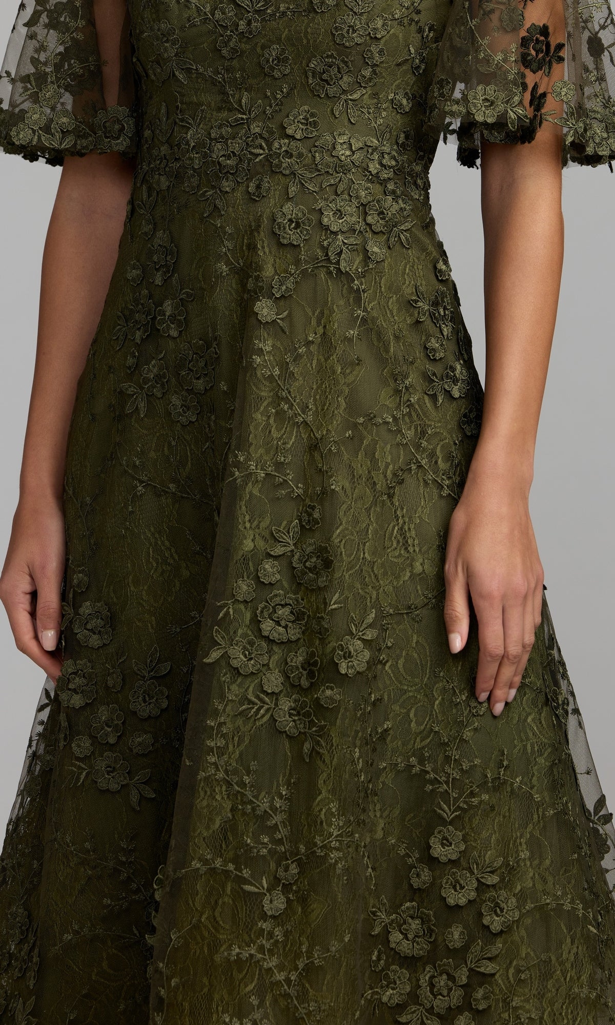 A woman models the Mac Duggal 69036 Lace Midi Wedding Guest Dress in olive green, showcasing its short sleeves and elegant floral lace with embroidered flower details—making it a chic choice for wedding guests. The image displays the dress from shoulders to mid-thigh.