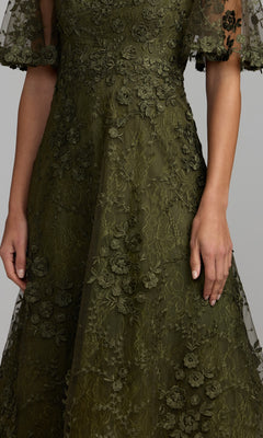 A woman models the Mac Duggal 69036 Lace Midi Wedding Guest Dress in olive green, showcasing its short sleeves and elegant floral lace with embroidered flower details—making it a chic choice for wedding guests. The image displays the dress from shoulders to mid-thigh.