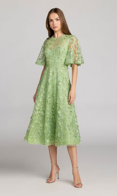 A woman wears the Mac Duggal 69036 Lace Midi Wedding Guest Dress, featuring light green lace with floral details and short sleeves, paired with open-toe heels against a plain gray background—perfect for wedding guests.