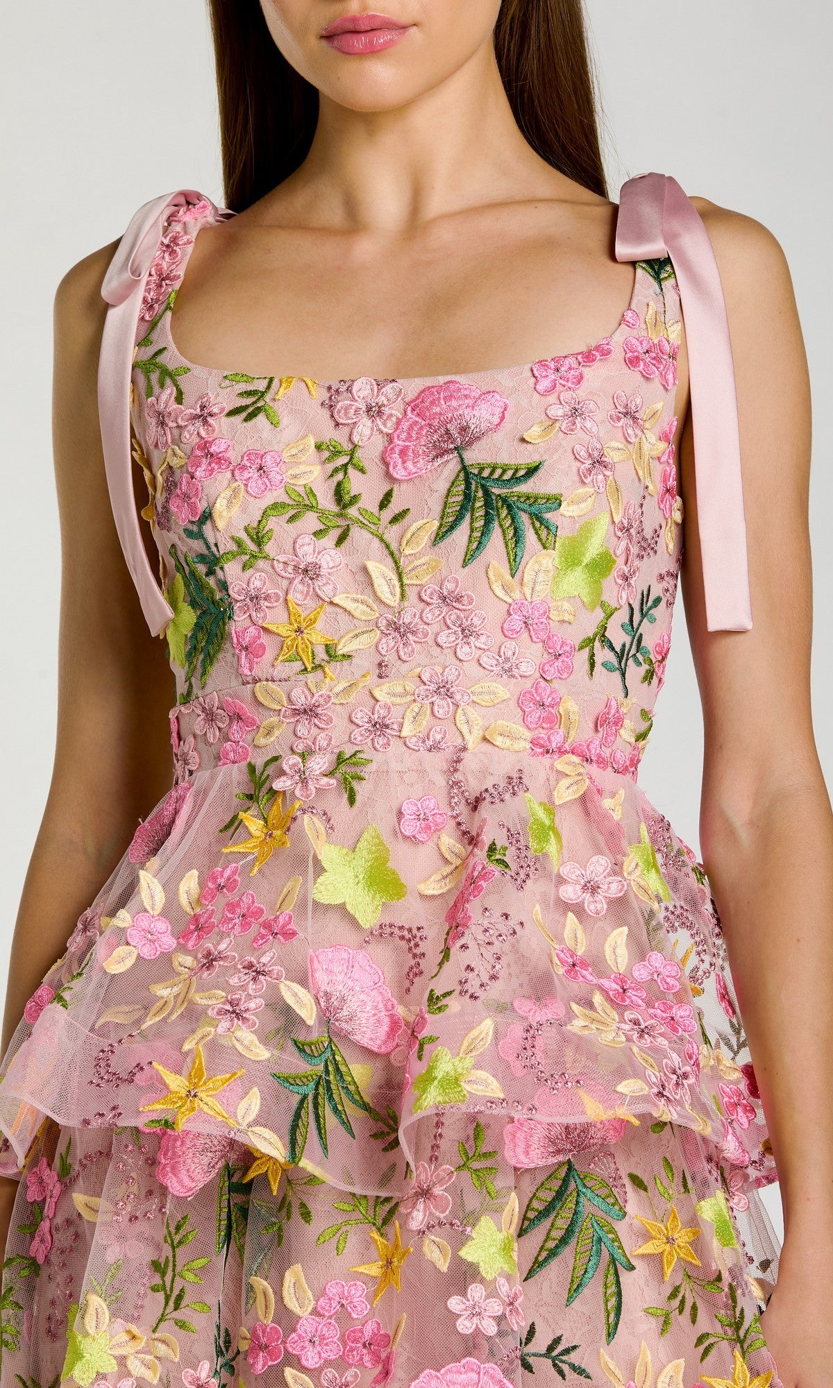 A woman wearing the Mac Duggal 69043 Midi Party Dress—a sleeveless pink floral design with delicate embroidery, shoulder bows, and a tiered A-line skirt—shown from the chest down.