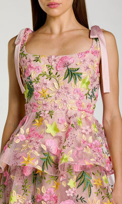 A woman wearing the Mac Duggal 69043 Midi Party Dress—a sleeveless pink floral design with delicate embroidery, shoulder bows, and a tiered A-line skirt—shown from the chest down.