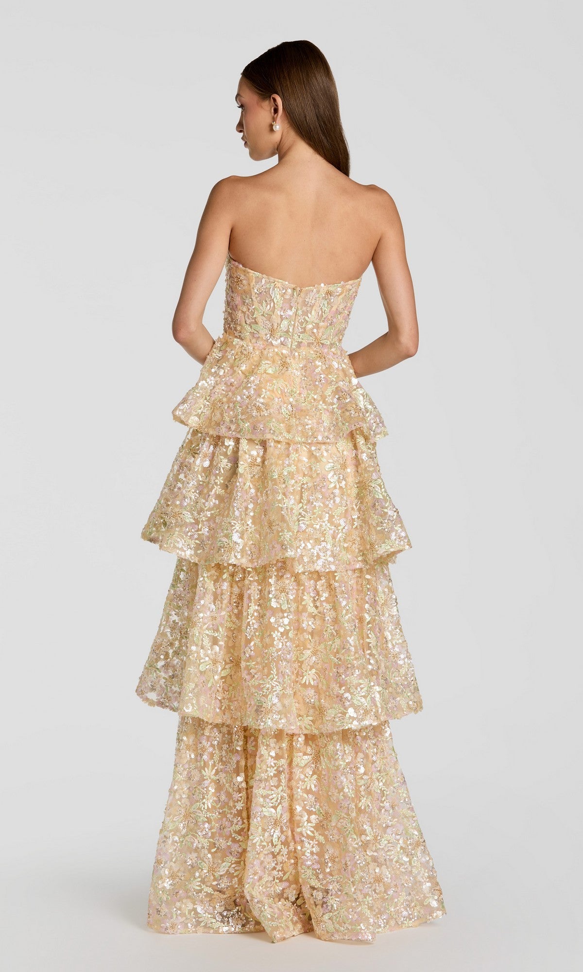 A woman facing away, with straight brown hair, wears the Mac Duggal 69044 Long Formal Dress—a strapless, tiered floor-length gown adorned with iridescent floral sequins—against a plain light background.