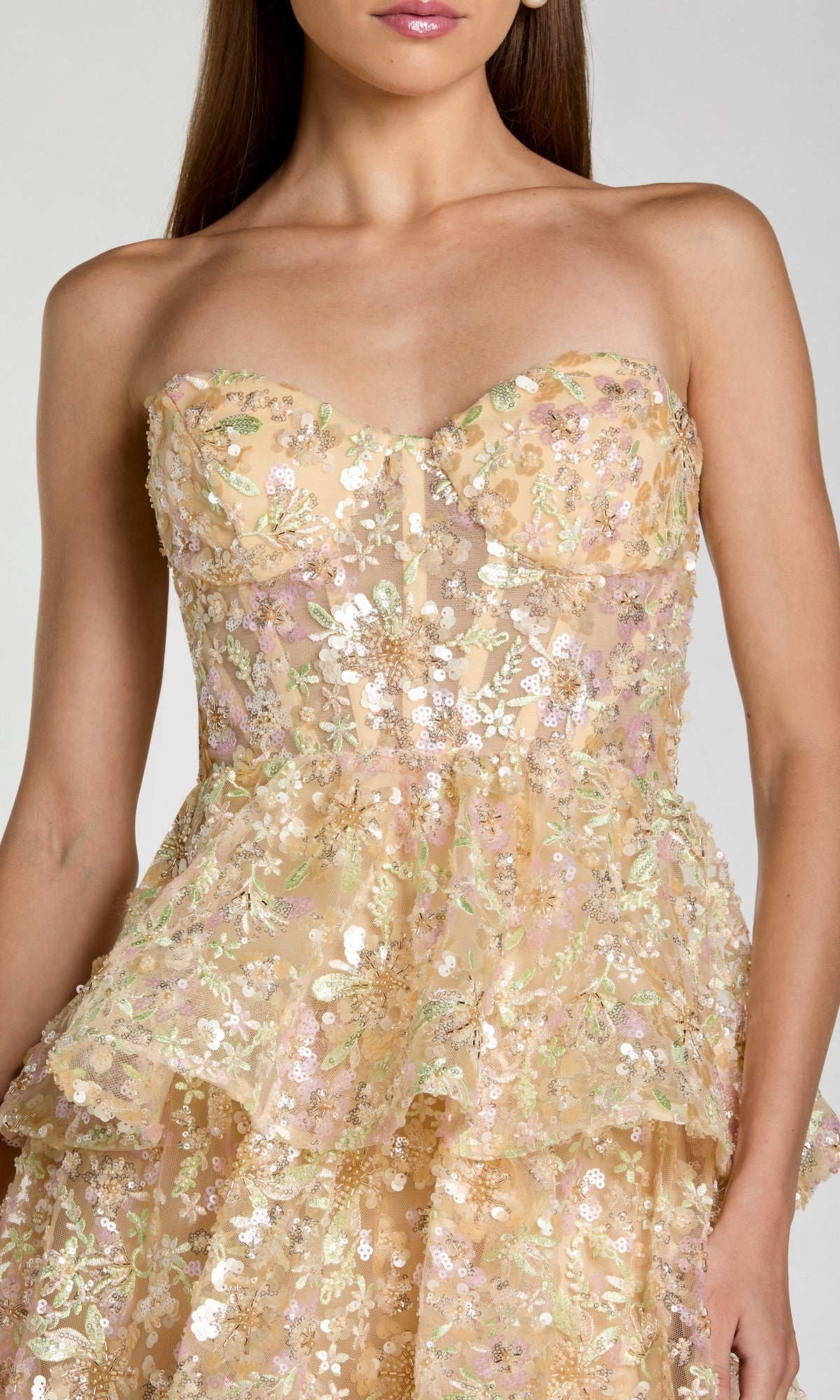 A woman wears the Mac Duggal 69044 long formal dress, a strapless gown in a soft peach tone with layered floral sequin embroidery and a sweetheart neckline. The ruffle-tiered A-line silhouette and exquisite detailing throughout create a romantic look.