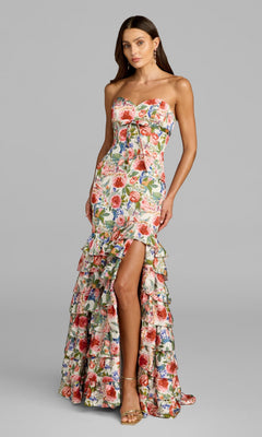 A woman is wearing the Mac Duggal 69059 long formal dress in Ivory Multi, which features a strapless floral design, mermaid silhouette with a front slit, and a ruffled hem, while standing against a plain gray background.