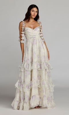 A woman stands against a plain background, wearing the Mac Duggal 69061 Long Formal Dress in Ivory Multi—a sleeveless, floor-length chiffon gown adorned with floral patterns, tiered ruffles, and delicate bow details on the straps for an effortlessly elegant A-line silhouette.