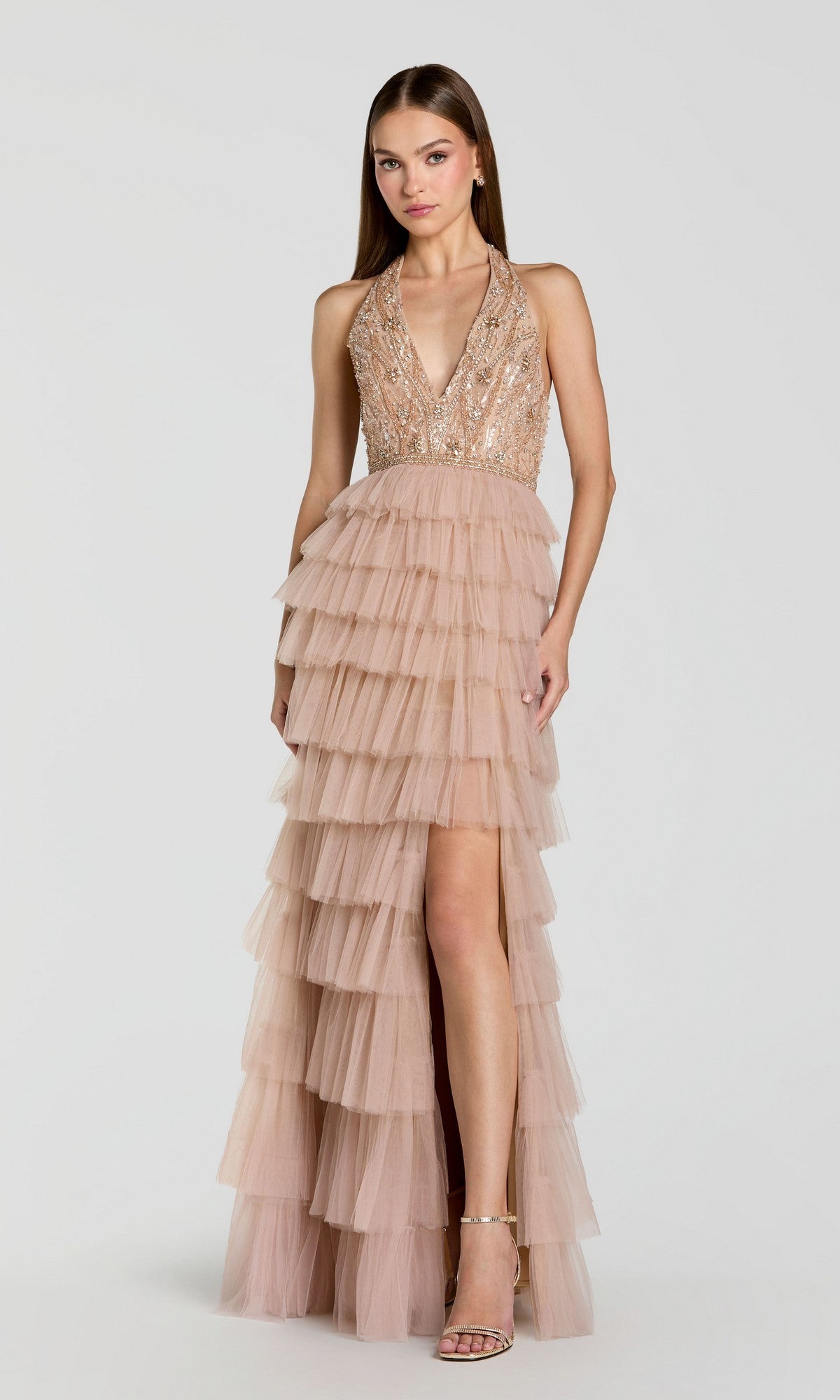 A woman wears the Mac Duggal 69062 Long Formal Dress—a sleeveless nude sequined halter gown featuring a tiered tulle skirt and high slit—while standing against a plain light background.