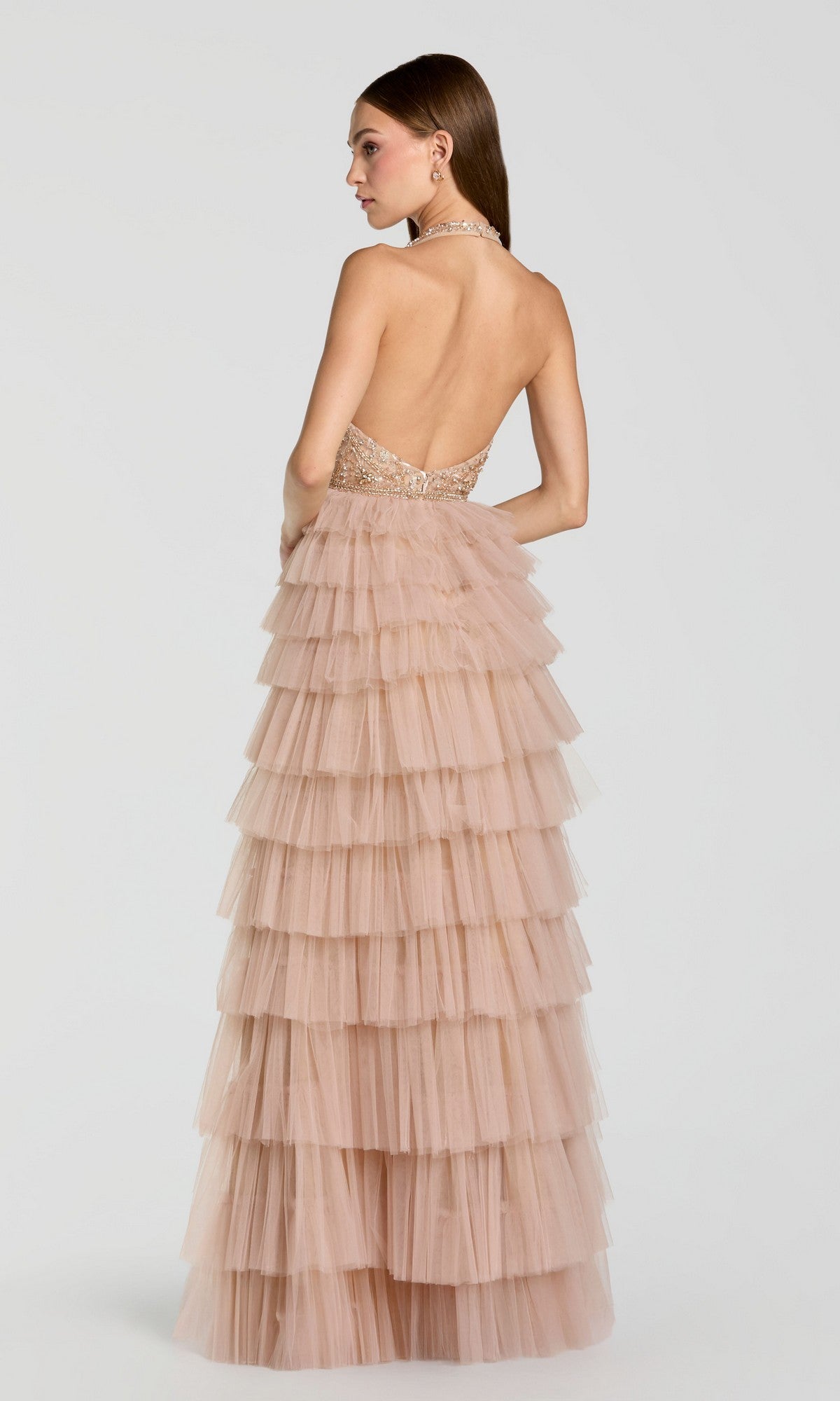 A woman stands against a plain light background, facing away from the camera, wearing the Mac Duggal 69062 long formal dress—a backless and strapless layered blush pink tulle gown with a fitted bodice.