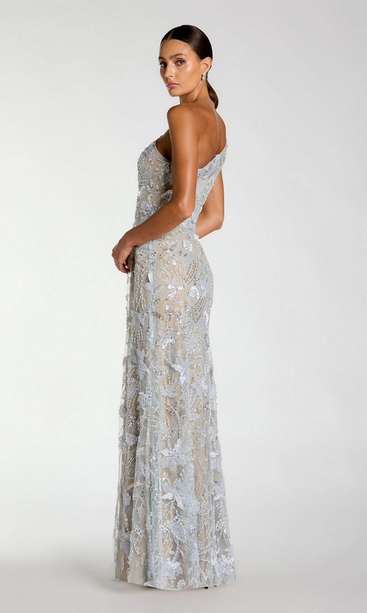 A woman stands in profile against a plain light background, wearing the Mac Duggal 69077 long formal dress—a sophisticated, light blue gown featuring floral lace embroidery, sequin embellishments, and a one-shoulder silhouette.