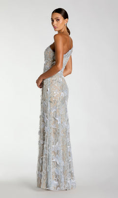 A woman stands in profile against a plain light background, wearing the Mac Duggal 69077 long formal dress—a sophisticated, light blue gown featuring floral lace embroidery, sequin embellishments, and a one-shoulder silhouette.