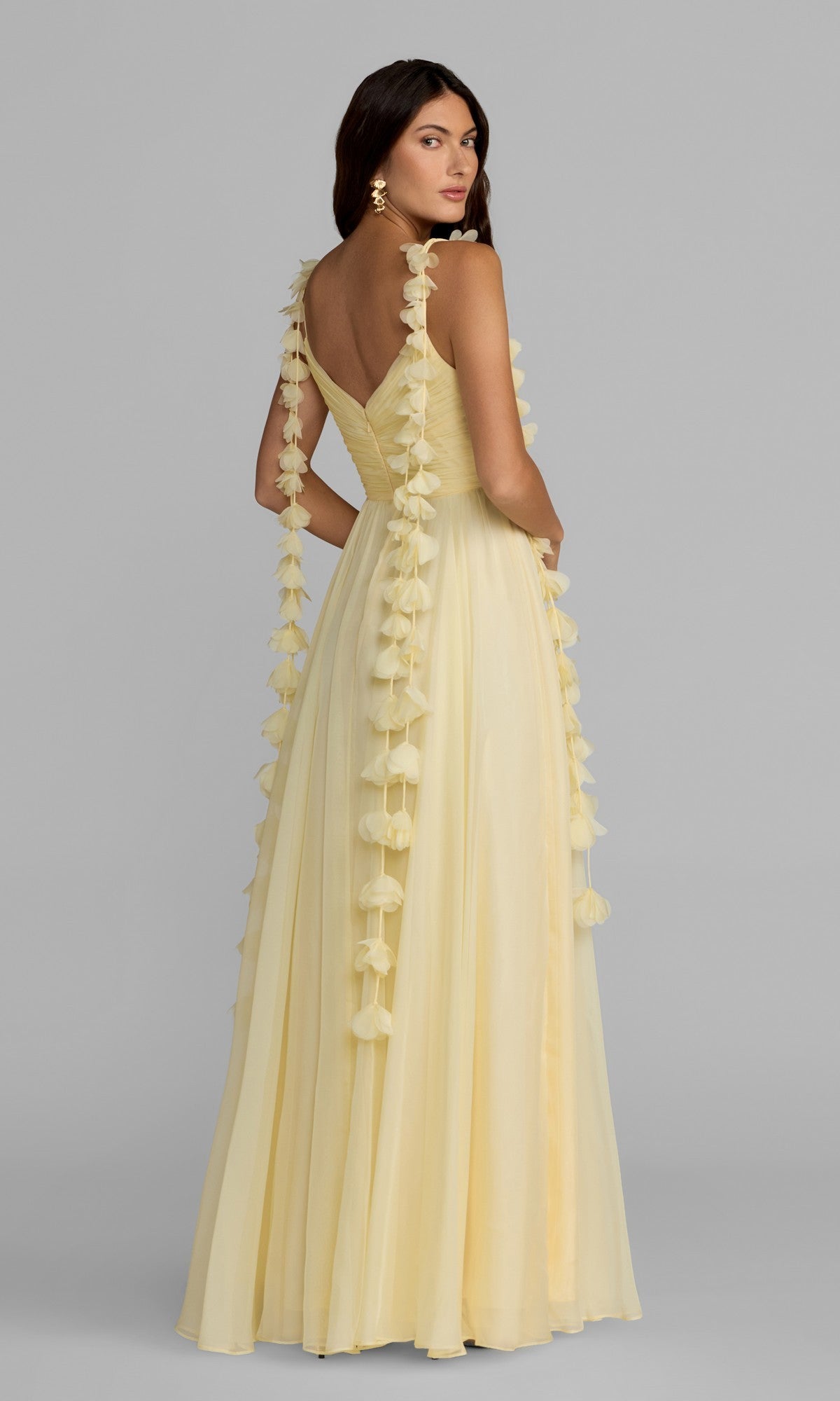 Wearing the Mac Duggal 69086 long formal dress—an A-line, pale yellow gown featuring floral detailing on the shoulder straps—a woman stands gracefully, looking over her shoulder against a plain gray background.