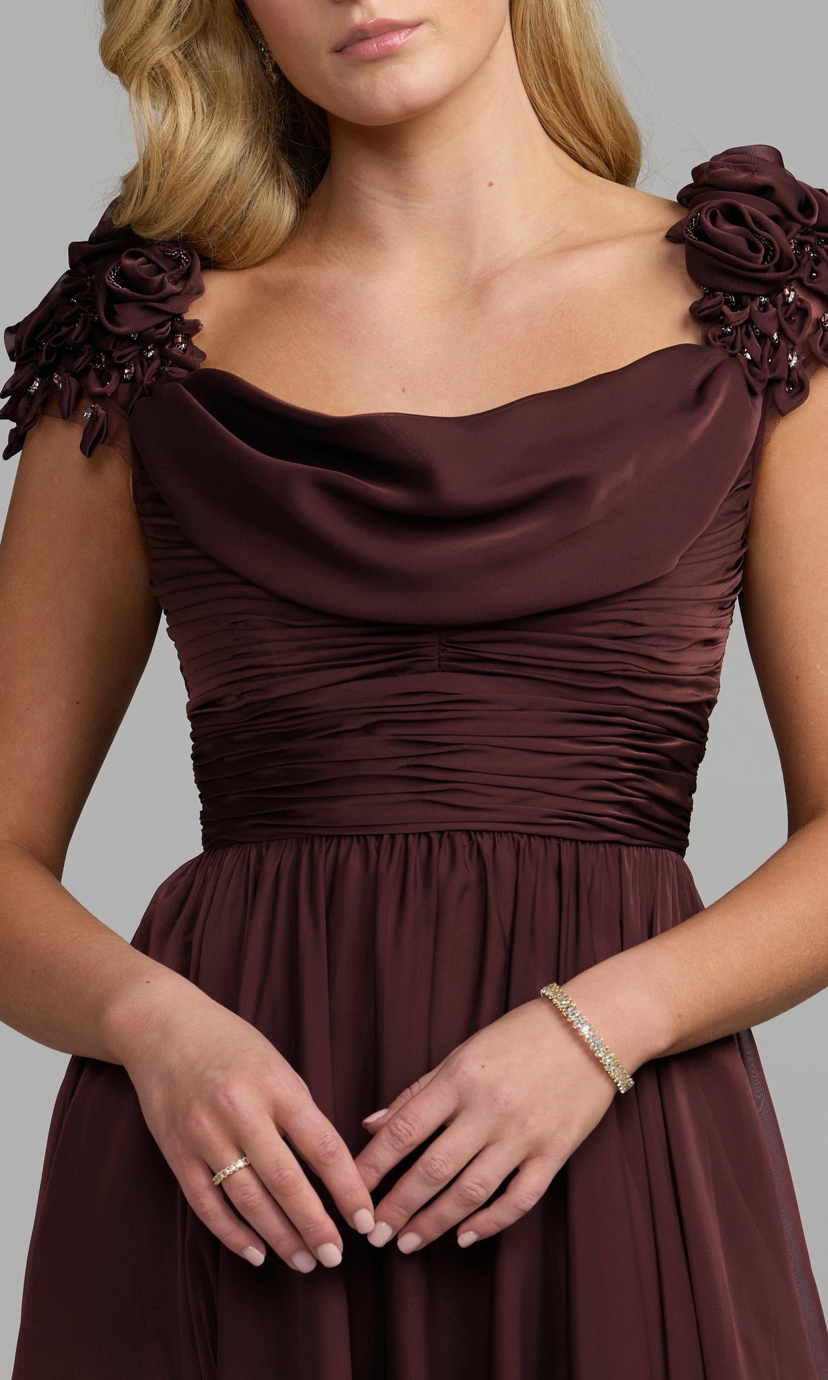 A woman stands against a neutral background wearing the Mac Duggal 69088, a sleeveless burgundy long chiffon formal dress featuring floral shoulder details and a subtle cowl neckline. She completes the elegant look with a bracelet and ring, capturing the timeless sophistication of Mac Duggal designs.