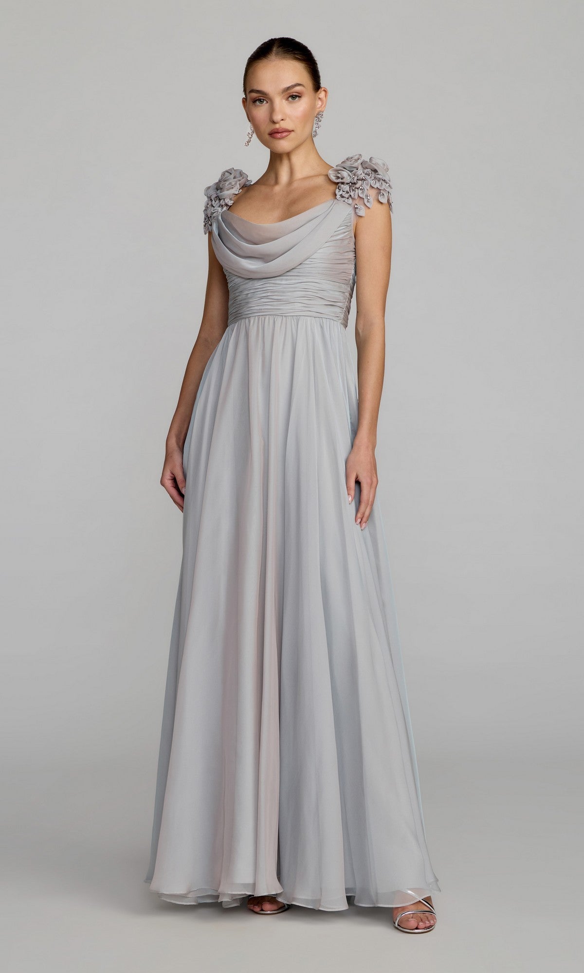 A woman stands against a plain light background wearing the Mac Duggal 69088 long formal dress in platinum—a light gray A-line gown featuring floral appliqué on the shoulders, a draped bodice, and open-toe heels.