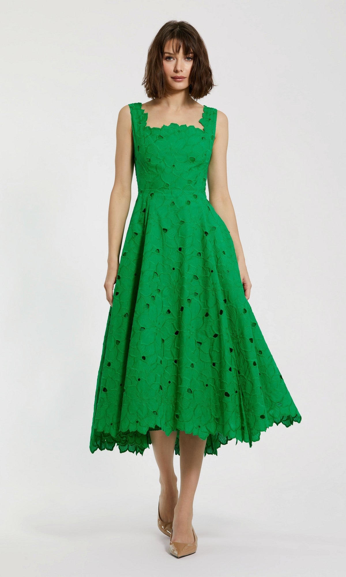 A woman stands against a plain white background wearing the Mac Duggal 69096 midi party dress in spring green—a sleeveless, eyelet-embroidered piece with a lace pattern and scalloped edges—paired with nude pointed-toe heels, adding an elegant yet playful touch to her look.