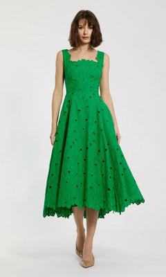 A woman stands against a plain white background wearing the Mac Duggal 69096 midi party dress in spring green—a sleeveless, eyelet-embroidered piece with a lace pattern and scalloped edges—paired with nude pointed-toe heels, adding an elegant yet playful touch to her look.