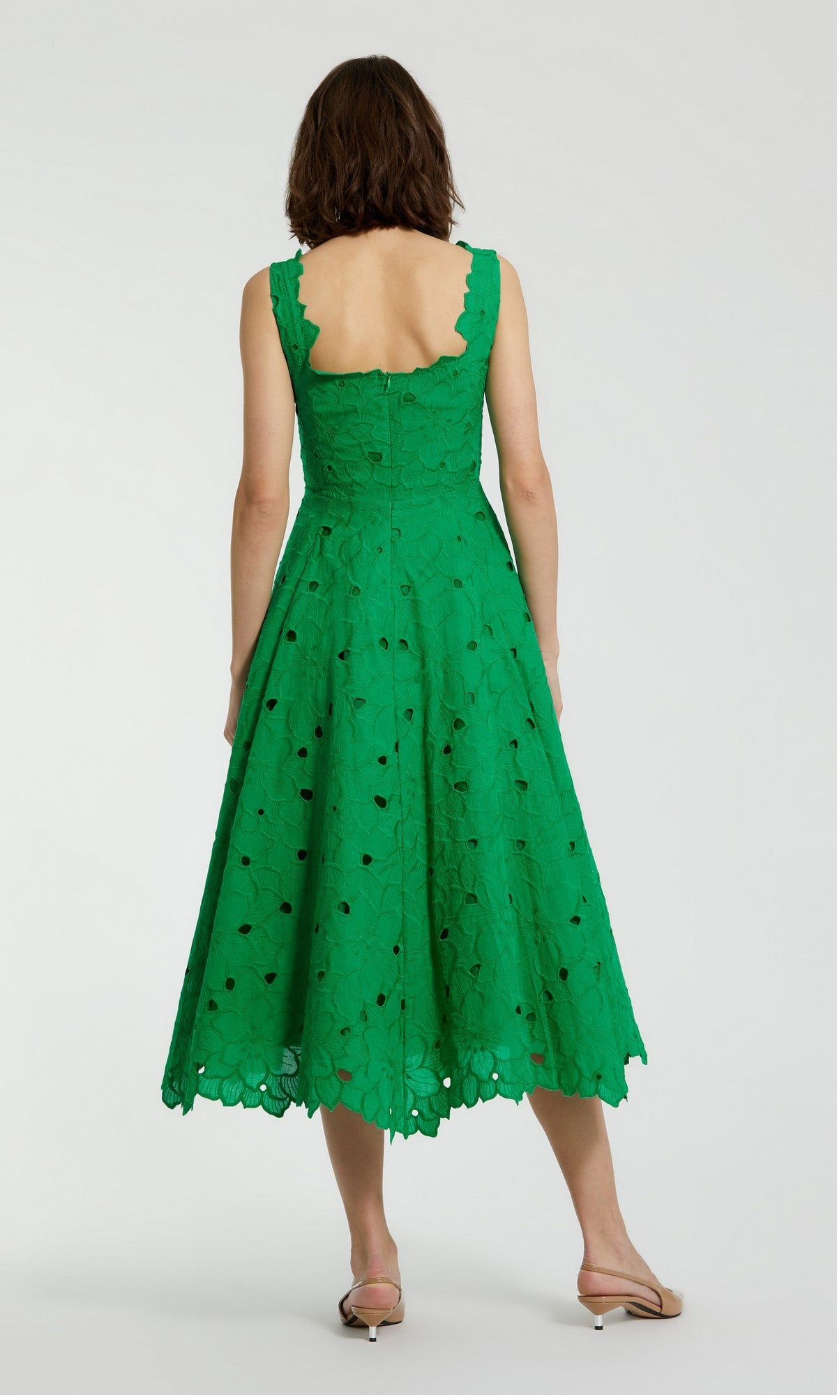 A woman stands with her back to the camera, wearing the Mac Duggal 69096 midi party dress in green—featuring a sleeveless design, scalloped hem, and open back—paired with light-colored flat sandals.