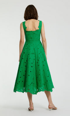 A woman stands with her back to the camera, wearing the Mac Duggal 69096 midi party dress in green—featuring a sleeveless design, scalloped hem, and open back—paired with light-colored flat sandals.