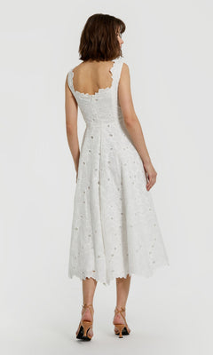 A woman stands facing away, displaying the elegant back of the Midi Party Dress: Mac Duggal 69096—a sleeveless white floral midi party dress adorned with delicate lace and eyelet-embroidered details—paired with open-toe heels.
