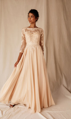 A woman poses before a neutral backdrop in the Eureka 6909 long-sleeve formal dress, showcasing its modest A-line silhouette with an ivory lace bodice and flowing chiffon skirt, holding one side of the gown.