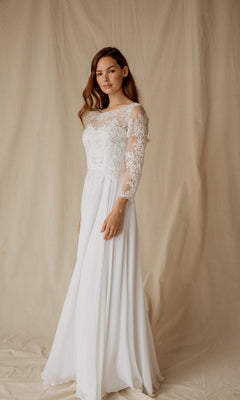 A woman models the Eureka 6909, a modest long-sleeve formal dress with lace accents, in front of a beige fabric backdrop—an elegant white gown ideal for special occasions.