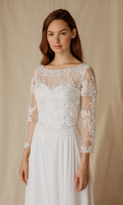 A woman stands before a beige backdrop in the Eureka 6909, a long formal dress featuring sheer long sleeves and delicate floral embroidery.