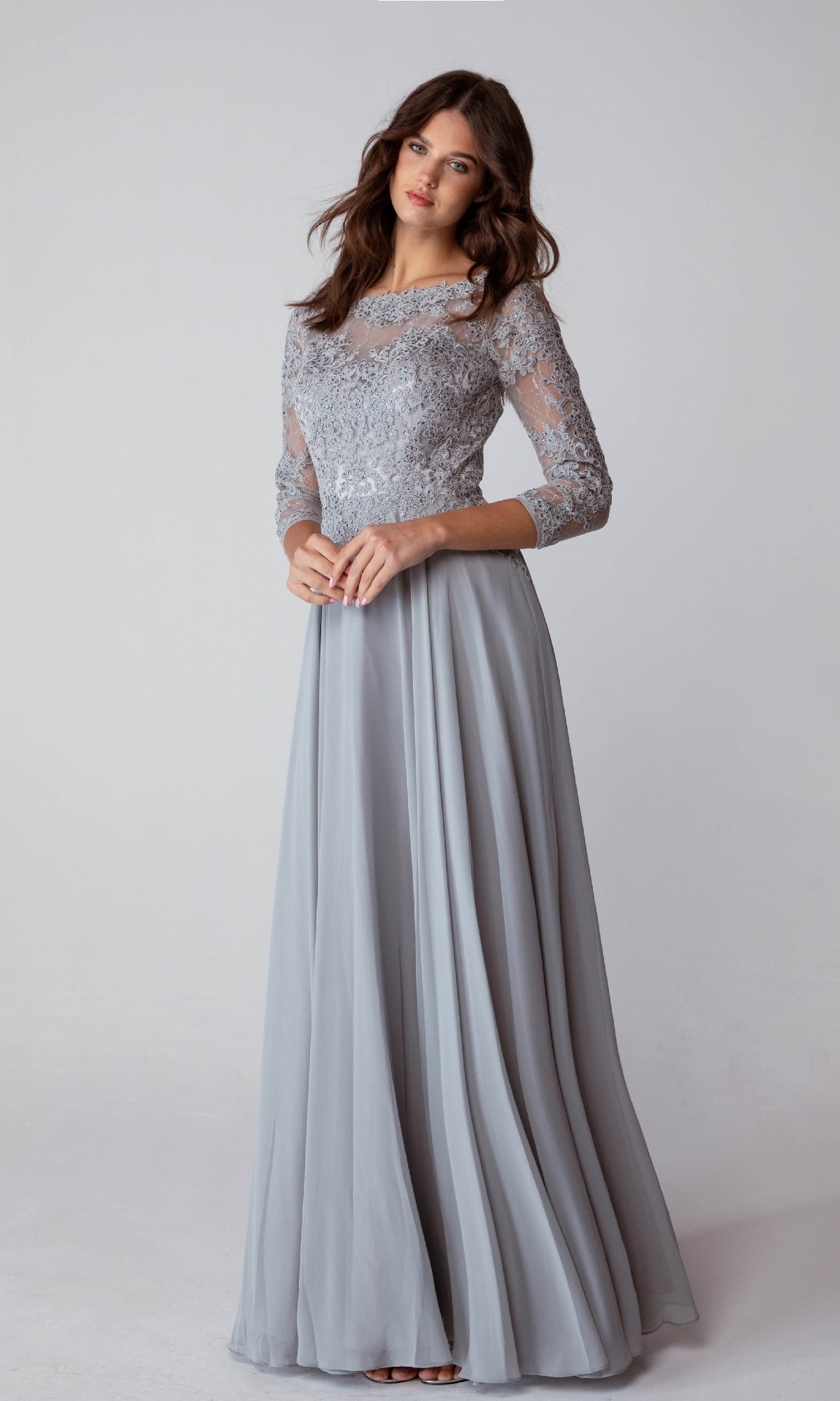 A woman stands against a plain background wearing the Eureka 6909, a long-sleeve, floor-length formal dress in light gray with a delicate lace bodice.