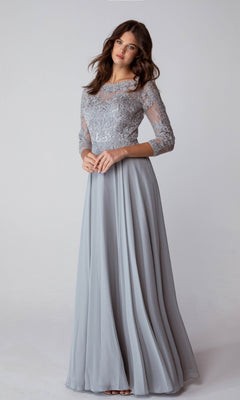 A woman stands against a plain background wearing the Eureka 6909, a long-sleeve, floor-length formal dress in light gray with a delicate lace bodice.