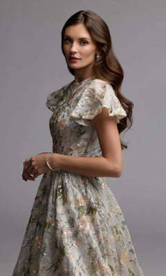 A woman stands with hands clasped, gazing at the camera while wearing the Mac Duggal 69107 Tea-Length Party Dress—featuring a sequin floral pattern and flutter sleeves—set against a plain gray background.