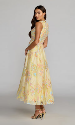 A woman stands in profile against a plain background, wearing the Tea-Length Party Dress: Mac Duggal 69111—an ankle-length, sleeveless yellow gown adorned with pastel floral embroidery—paired with gold heels and drop earrings.