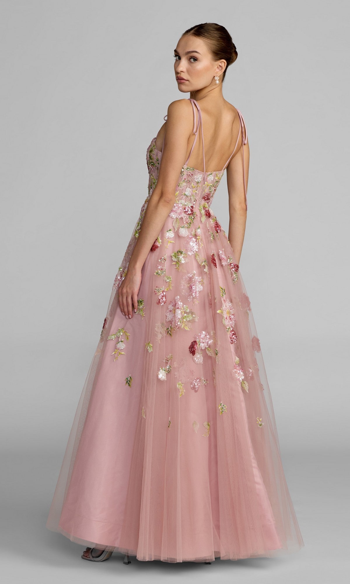 A woman in the Mac Duggal 69143 Long Formal Dress, an elegant pink A-line gown with delicate floral embroidery and thin straps, stands facing slightly away while looking over her shoulder.
