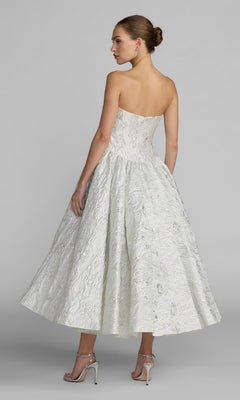 A woman in the Mac Duggal 69146 tea-length party dress, featuring a textured white brocade fabric and a strapless sweetheart bodice, stands with her back to the camera, looking over her shoulder while wearing silver heels against a plain gray background.