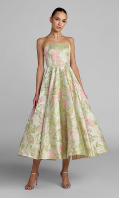 A woman poses in the Tea-Length Party Dress: Mac Duggal 69147, a strapless sage multi floral brocade dress in pastel green and pink tones, paired with silver high-heeled sandals against a plain grey background.