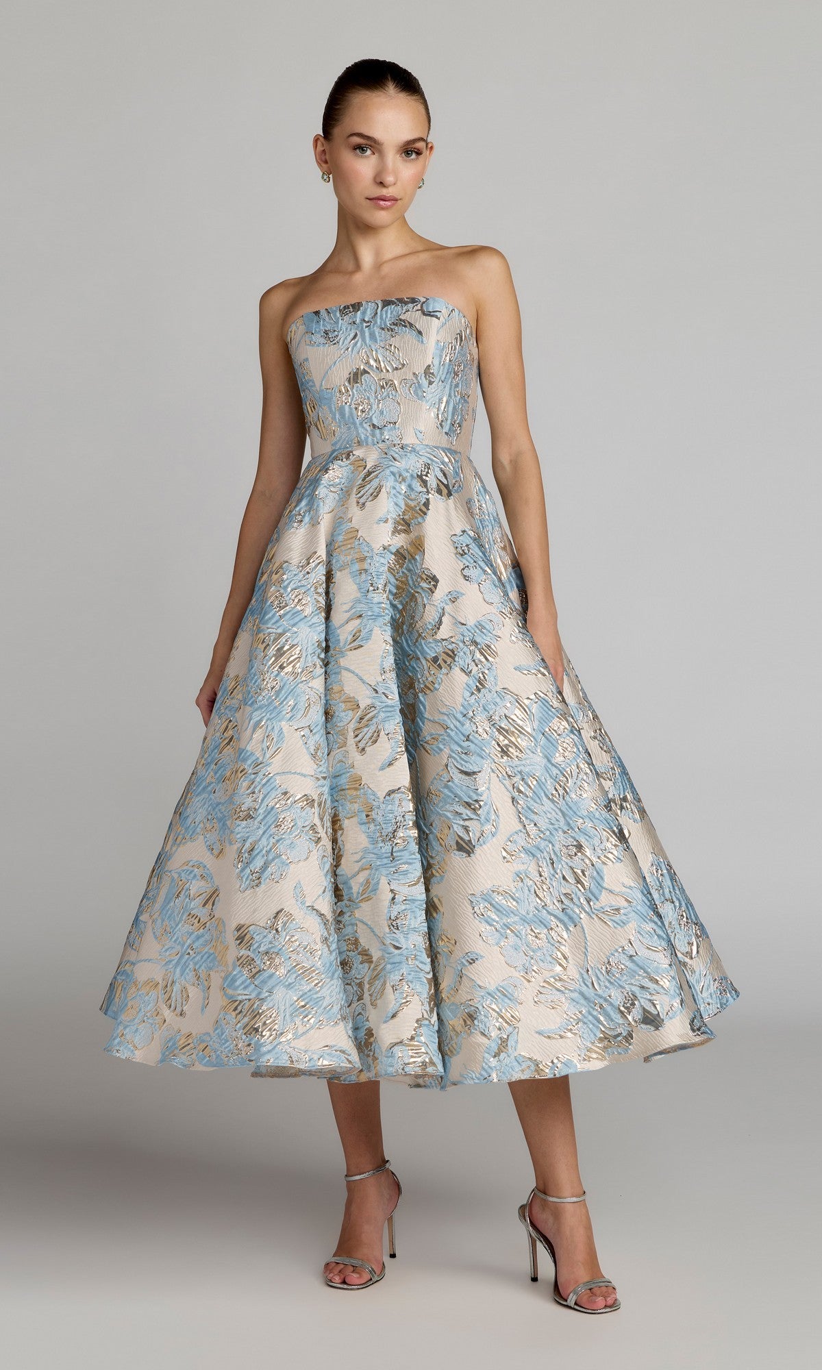 A woman wearing a strapless, powder blue tea-length party dress with a full skirt—Mac Duggal 69149—paired with silver high-heeled sandals, stands against a plain background.