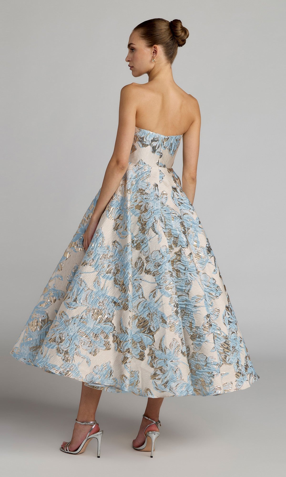 A woman stands against a plain grey background wearing the Tea-Length Party Dress: Mac Duggal 69149—a strapless, floral brocade tea-length dress featuring blue and gold patterns—paired with silver high heels and her hair styled in a neat bun.