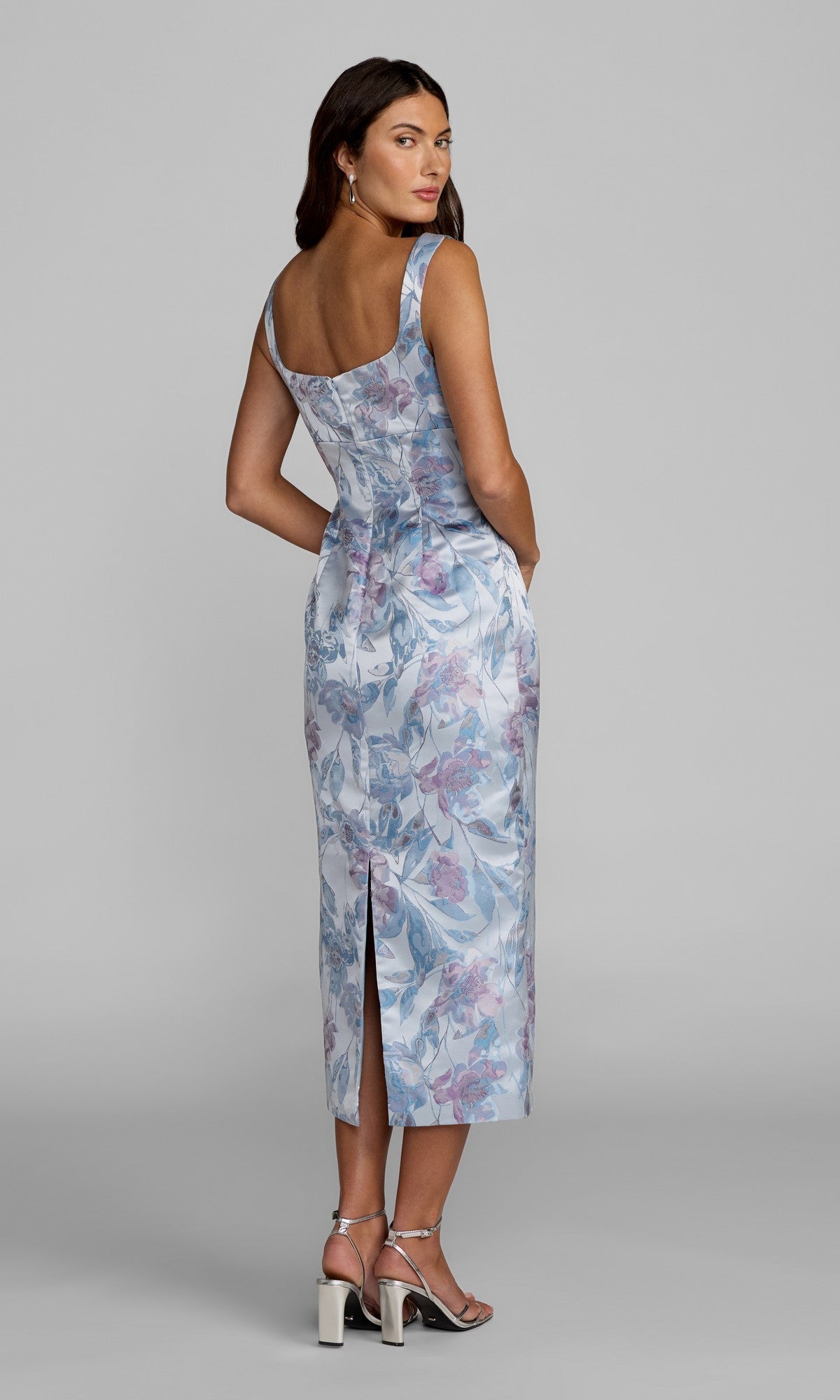 A woman poses against a plain gray background, looking over her shoulder while wearing the Floral Midi Wedding Guest Dress: Mac Duggal 69152—a light blue floral midi dress featuring a sleeveless design and back slit—offering an elegant, chic option for any wedding guest.