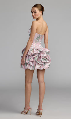 A woman wearing the Mac Duggal 69156 short party dress, featuring a strapless bodice and pastel-patterned ruffled skirt, stands in high heels and looks over her shoulder against a plain light gray background.